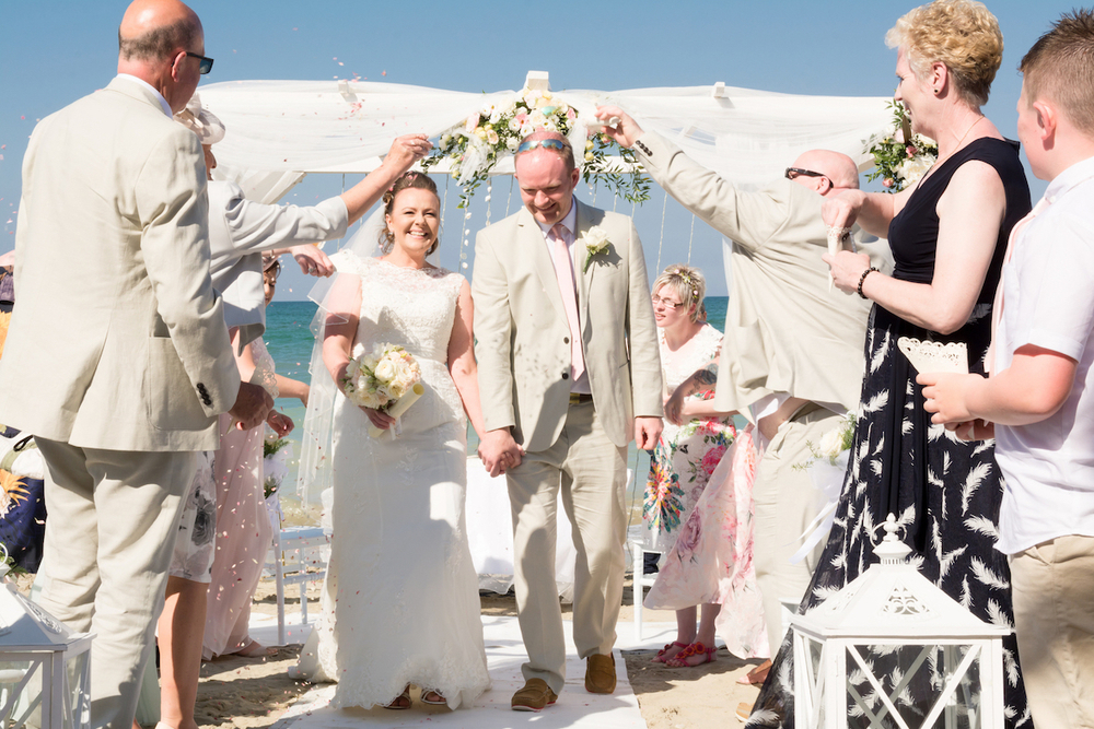 bride and groom walking along an aisle on a beach with wedding guests throwing confetti