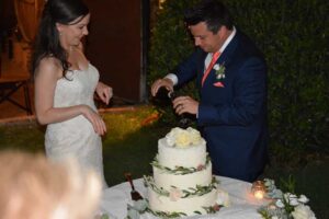just married groom pours a glass of Prosecco for his new wife in front of a decorated round table with a three tier white iced wedding cake on it