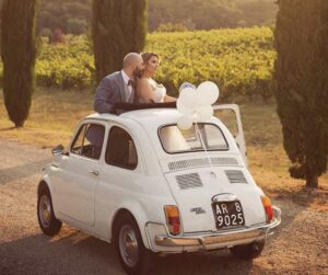 newly married couple standing up out of the sun roof in a white Fiat 500 parked next to a vineyard