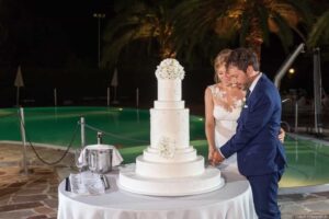 a newly married couple in a garden in front of a swimming pool make the first cut of a large Italian wedding cake