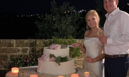 Happy bride and groom cut the cake on an open air terrace in Italy with a view of hill top town behind them.