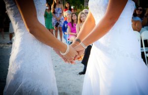 Gay couple hold hands at their wedding ceremony on a sandy beach.