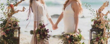 Two women hold hands and look into each other's eyes as they marry in a gay beach wedding. The sea is behind them and they are standing on the sand. There are floral compositions to each side of them creating a wedding alter type space.