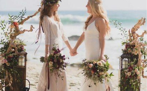 Two women hold hands and look into each other's eyes as they marry in a gay beach wedding. The sea is behind them and they are standing on the sand. There are floral compositions to each side of them creating a wedding alter type space.