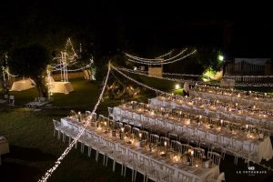 Long Italian dining tables set up for a wedding banquet. In a garden at night, obviously warm and with fairy lights overhead.