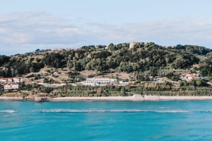 Italian wedding venue on the sea shore photographed from the sea. You can see wooded hillsides behind the white structure and an abbey amongst the trees.