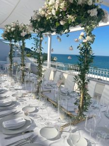 A beautiful wedding banquet table set up on a terrace above the sea. There is a canopy to keep the sun off the diners and flowers decorating the canopy.